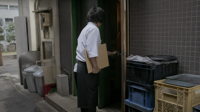資料を持って店舗の裏口に入る飲食店の運営者